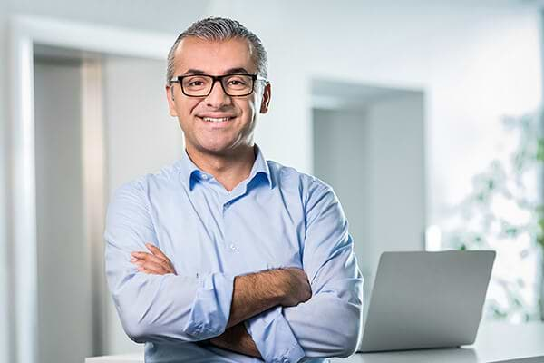 Mann mit schwarzer Brille, der die Arme verschränkt und in Kamera lächelt mit silbernem Notebook im Hintergrund