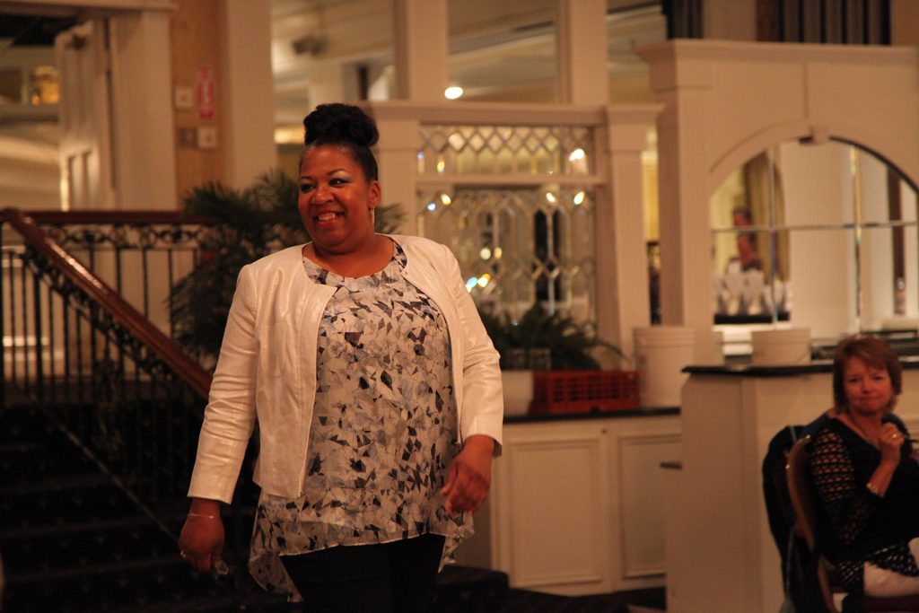 A woman wearing a white jacket and blue/white patterened shirt smiles while walking the runway