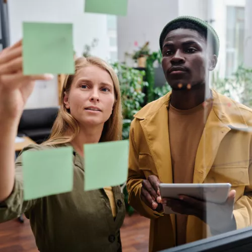 Two coworkers collaborating at a board covered in sticky notes, engaged in a productive brainstorming or planning session