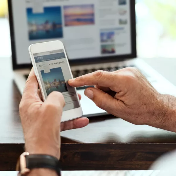 A close up shot of the hands of a person holing and taping their phone with their opened laptop in the background