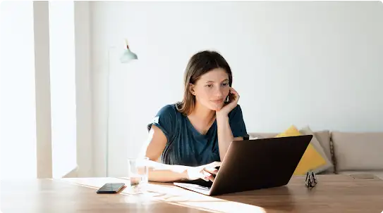 Une femme portant un haut bleu foncé utilise un ordinateur portable sur une table en bois. Un smartphone, un verre d’eau et une paire de lunettes sont également posés sur la table.