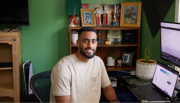 Um homem negro est� sentado em um home office sorrindo e olhando para a c�mera � sua direita. Ele tem cabelo curto e uma camiseta bege.