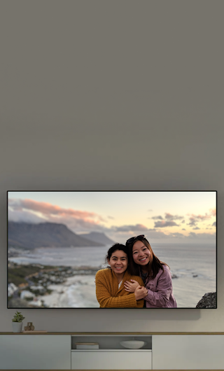 A TV in a living room showing a picture of two people standing with the ocean in the background. A phone with the same photo is in front of the TV.