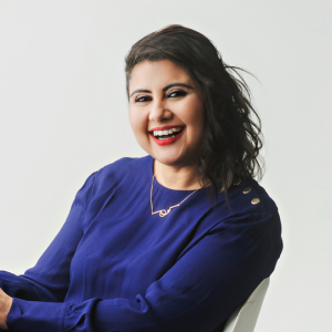 Portrait of Komal Singh, an Engineering Program Manager on the Google MUSE team. Smiling and sitting in a chair, she is turned to her left to face the camera and is wearing a blue outfit.