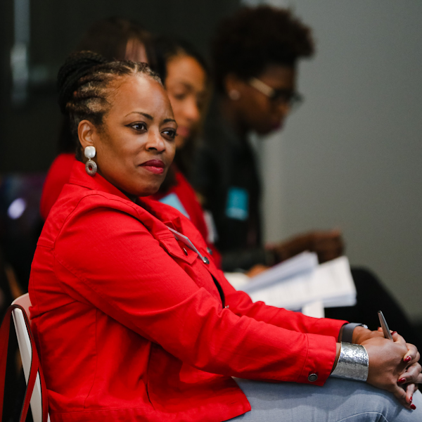 Black woman wearing a red jacket and chunky earrings sits next to other audience members, listening attentively