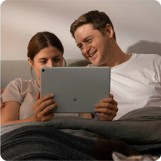 Dos personas descansando en una cama y sonriendo mientras ven contenido de entretenimiento en su tablet.