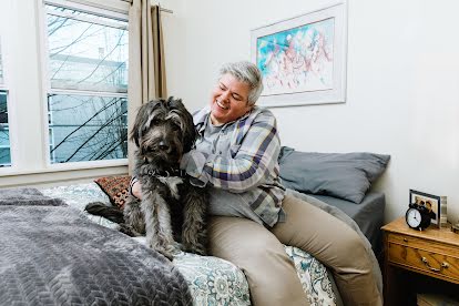 A smiling woman sits on a bed, her device-free zone, and holds a large grey shaggy dog.