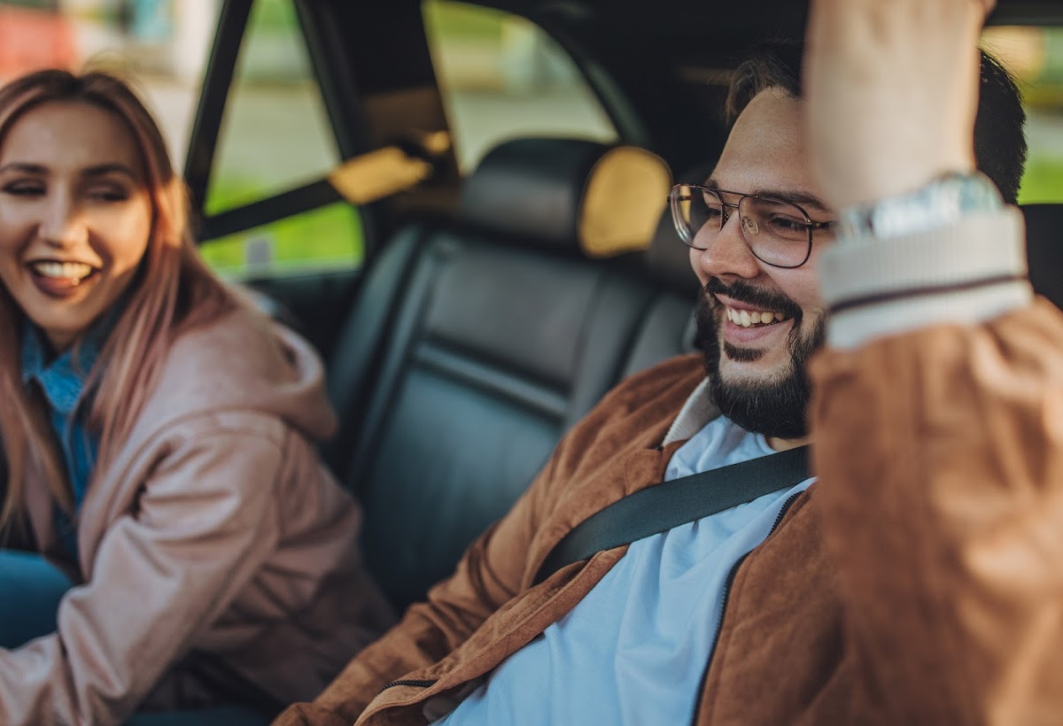 Looking through the drivers side window a bearded man with glasses grins widely, a women in the passengers seat is smiling while leaning forward.