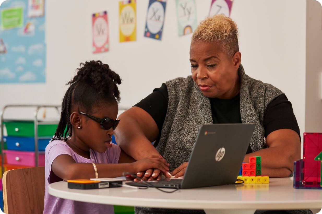 Una alumna joven con discapacidad visual utiliza un Chromebook con la ayuda de un lector de braille mientras la profesora, sentada a su lado, gu�a su mano.