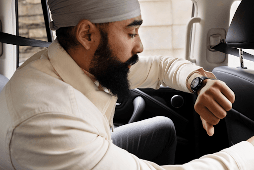 Um homem est� no banco de tr�s de um carro com o bra�o esquerdo pr�ximo ao rosto enquanto confere uma notifica��o no smartwatch. Ele est� inclinado para frente e segurando o encosto do banco do motorista, parecendo estar com pressa.