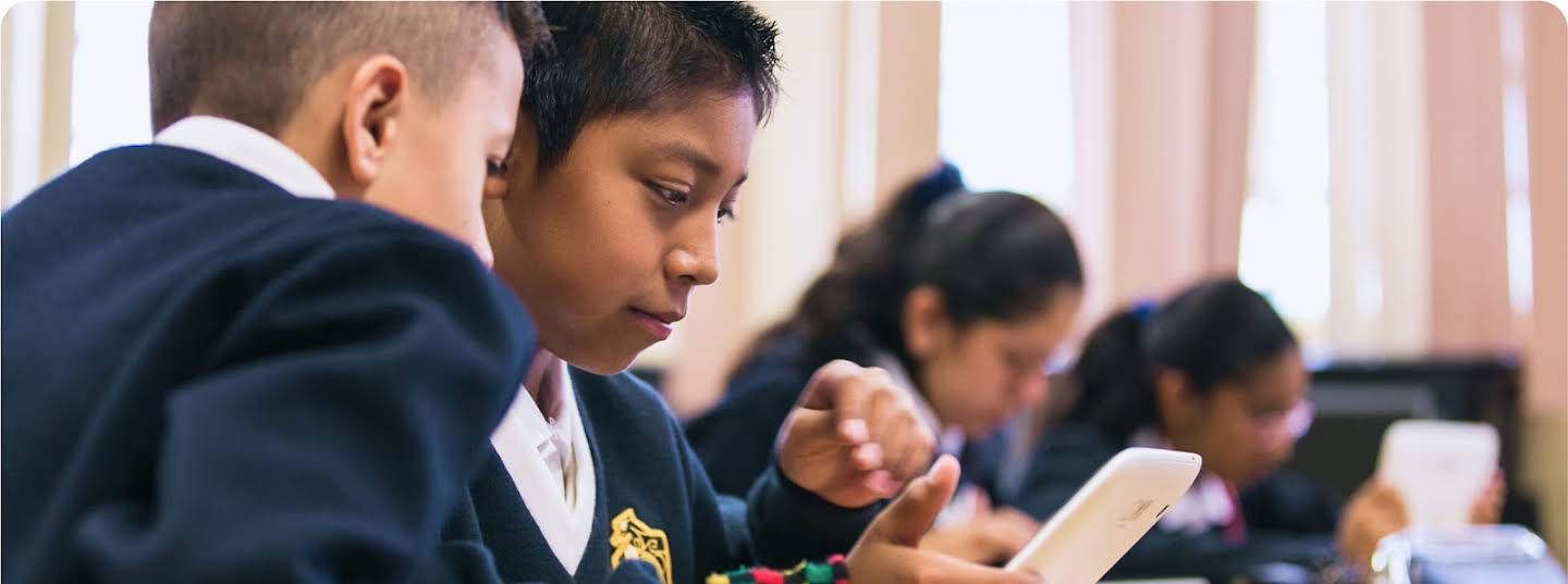 Two students look at the same tablet. In the background, two other students are working on tablets.