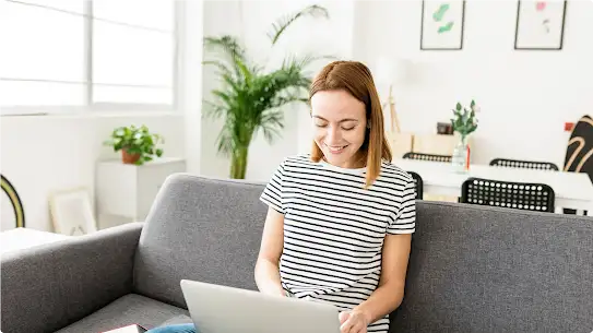 Une jeune femme portant un haut rayé noir et blanc est assise sur un canapé gris, avec un ordinateur portable ouvert sur les genoux. Elle tape sur les touches du clavier en souriant.