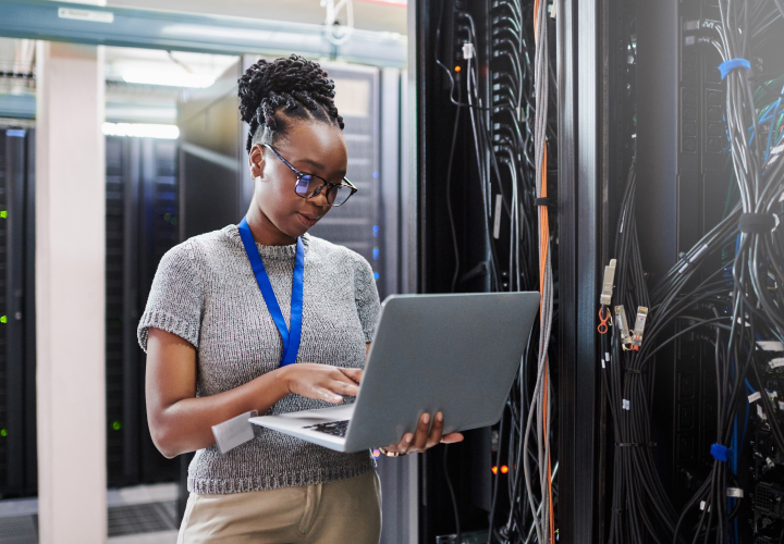 Eine Frau steht im Serverraum und sieht auf ihren Laptop.