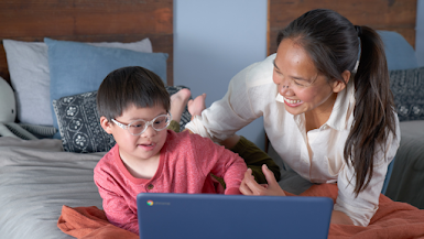 Young boy with a red long sleeve shirt and thick glasses lies on a bed staring at a Chromebook laptop, his mother lies smiling next to him