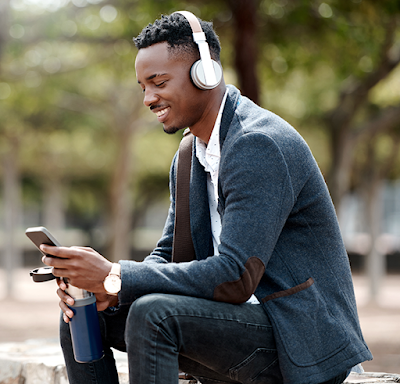 Un homme noir, professionnel, portant un casque d'�coute, sourit tout en tenant son appareil mobile et une bouteille d'eau alors qu'il est assis � l'ext�rieur sur un banc public.