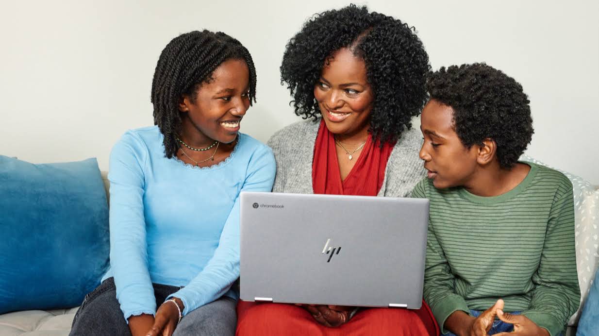 A mom typing on a Chromebook on the couch with two kids by her side.