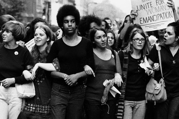 A crowd of women walk with their arms linked, chanting, and carrying signs at a women’s rights rally