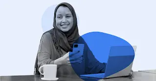 O femeie care poartă hijab zâmbește în timp ce folosește smartphone-ul și laptopul.