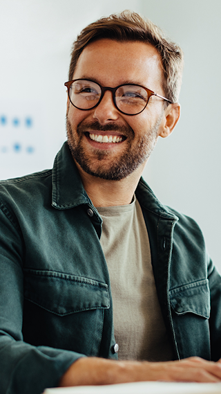 Un homme blanc � lunettes porte un t-shirt beige sous une veste verte, sourit et travaille sur son ordinateur portable. Il est assis dans une salle de conf�rence � c�t� de sa coll�gue asiatique qui porte un col roul� blanc et des lunettes.