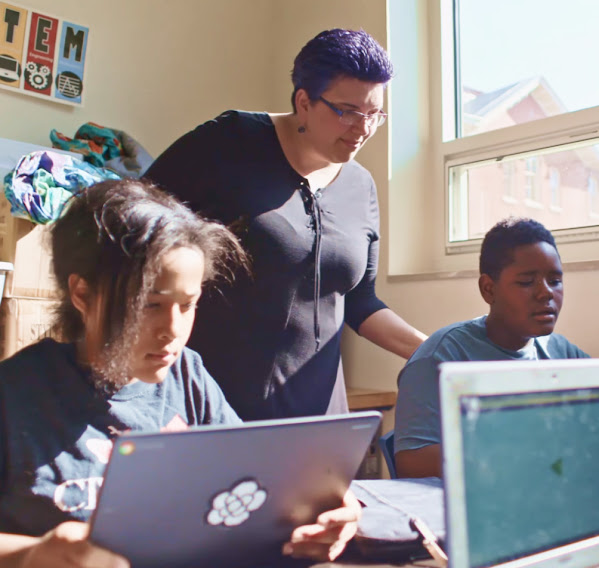 A teacher watches over two students while they work on laptops.