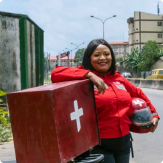 A woman wearing a red jacket stands on a sidewalk leaning against a red motorbike, holding a helmet.