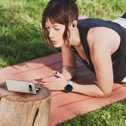 Persona haciendo ejercicio sobre una estera de yoga con un reloj inteligente Wears OS y auriculares, y mirando un tel�fono Android plegable.