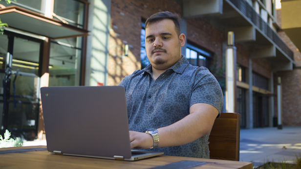 A man wearing a blue patterned button-down shirt sits and types on a laptop