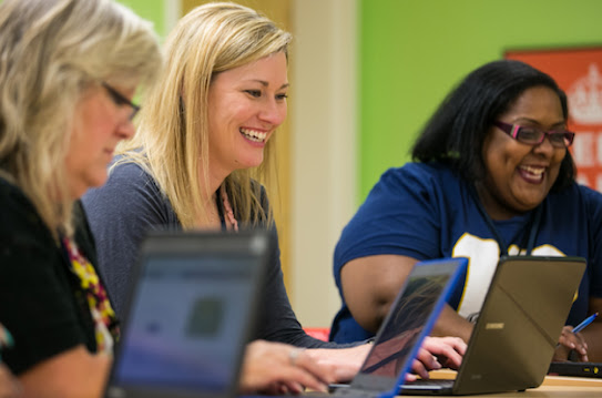 Three adult women sit at a long table with their laptops, laughing.