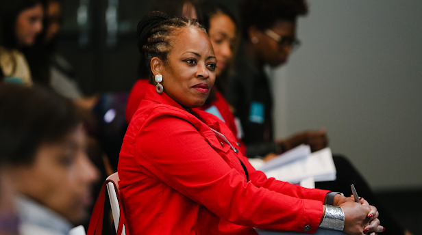 Black woman wearing a red jacket and chunky earrings sits next to other audience members, listening attentively