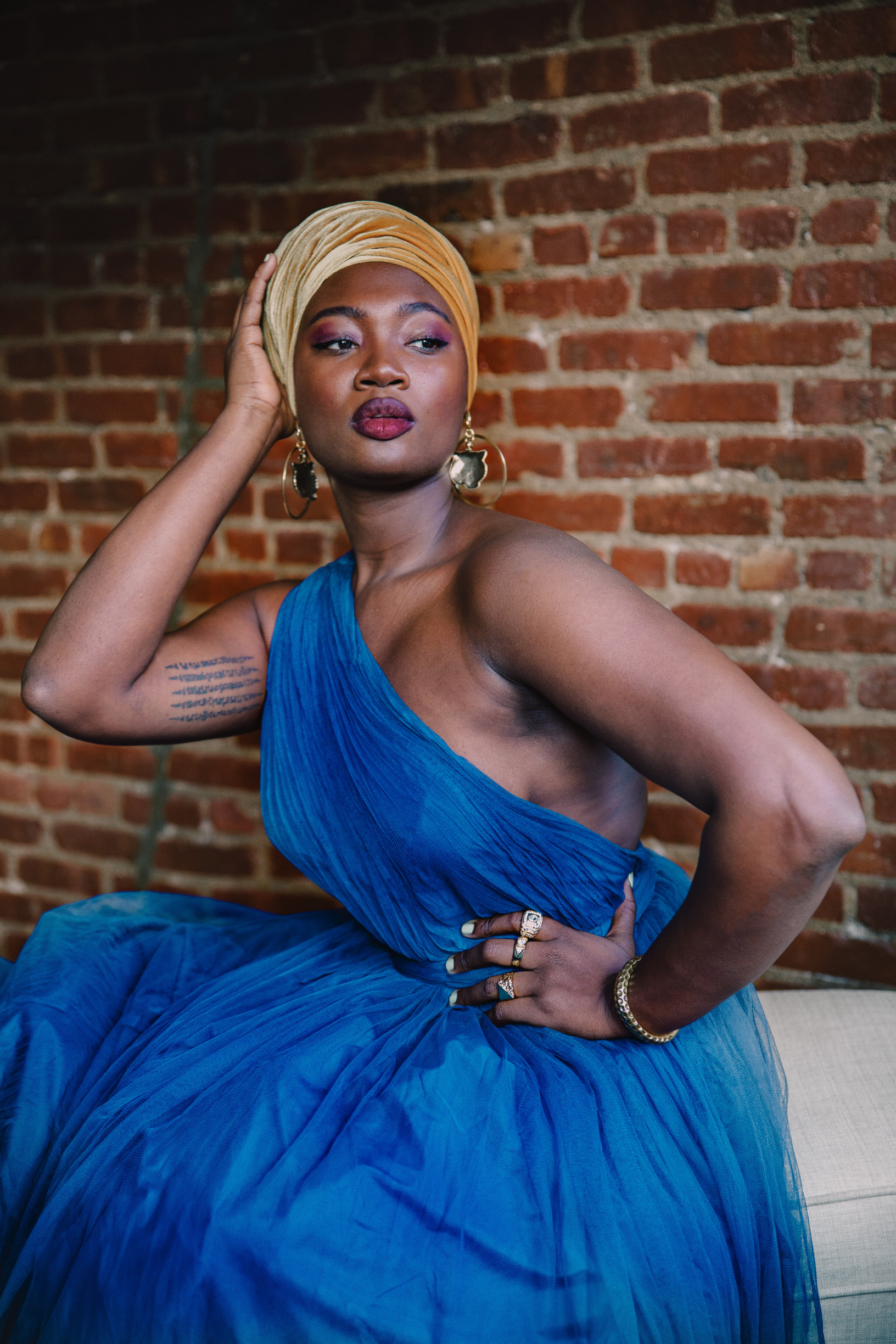 Solo photograph of Mama Cax sitting in a blue dress and yellow head scarf with her hand posed on her head