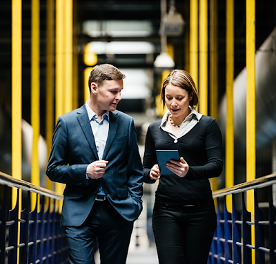 Een witte man met een blauw pak en een witte vrouw met een zwarte trui lopen door een datacenter, terwijl ze gegevens op een tablet bestuderen.