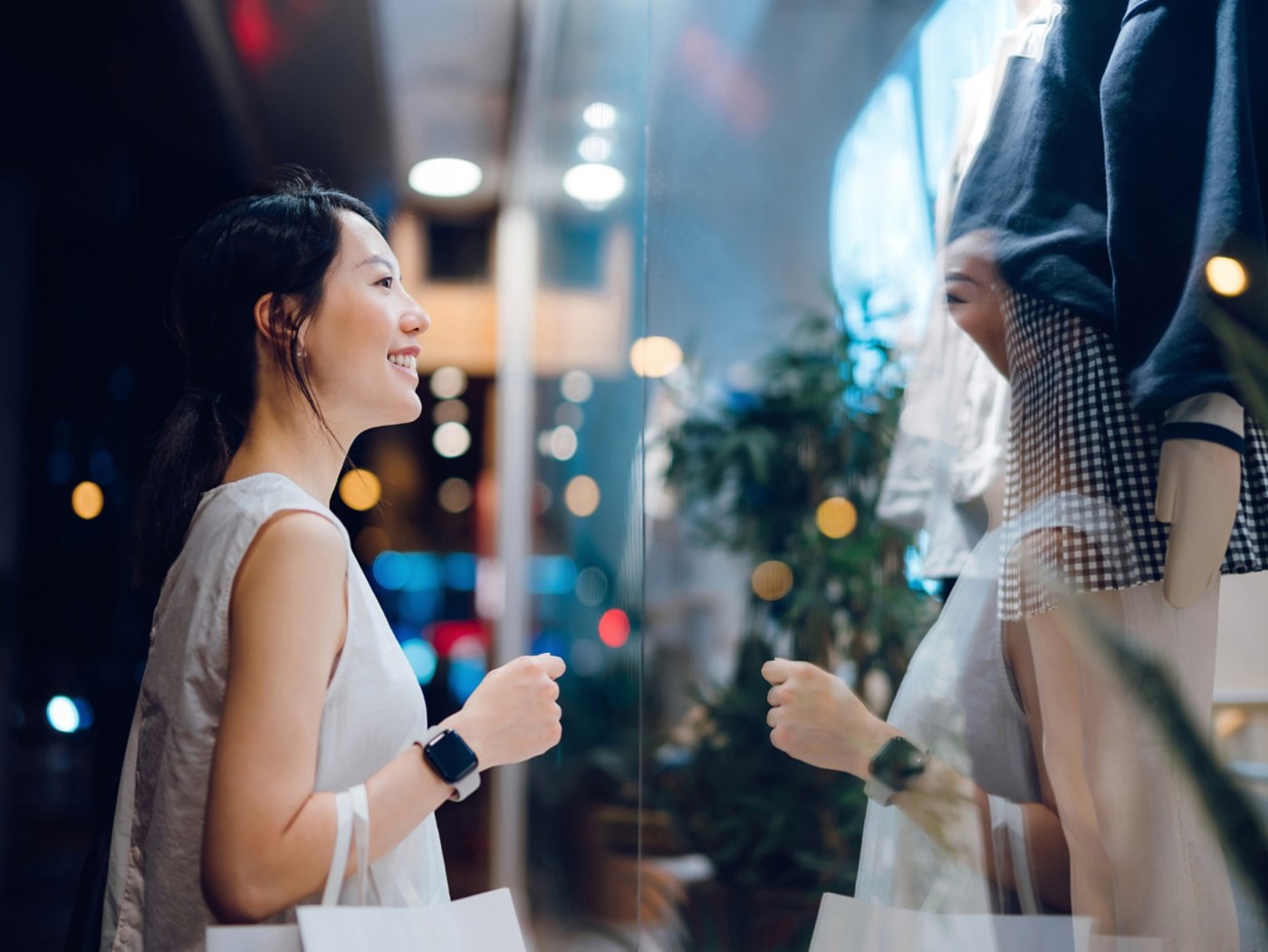 Smiling young Asian woman carrying a paper shopping bag, standing outside a boutique looking at shop window in the evening in city