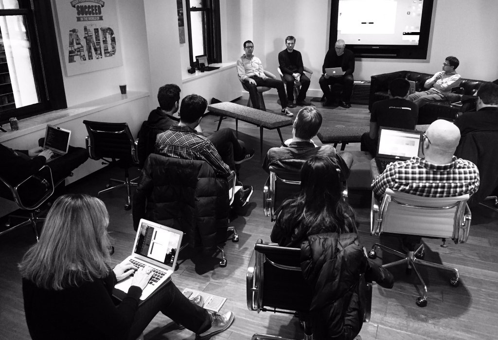 A black and white photo from a meetup. I am sitting with two other people next to the speaker, across from a small crowd.