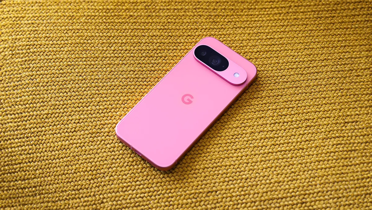 A pink Pixel 9 phone slightly tilted to the right, lying screen-down on a yellow knit fabric. The phone’s back is visible, showing its pill-shaped Camera Bar at the top of the device and a slightly darker shaded pink G in the center of the phone.