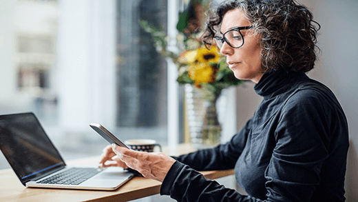 Mature businesswoman sitting at cafe looking at her cell phone while working on laptop computer. Woman reading text message at coffee shop.