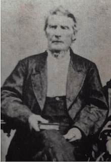 An elderly man is seated with a book in his right hand in his lap