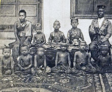 Sepia tinted photo of seated woman and older man separated by three children sitting on a bench, with several children sitting on the floor in front of them