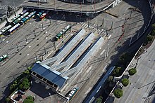 A parking lot filled with buses, seen from above. A station with several blue-roofed platforms can be seen towards the bottom, with lanes for buses and tracks for trains.