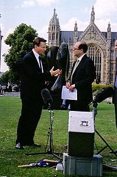 Two men in suits, stood on a grassy field in front of a Gothic style building. There is a tree on the left side, and microphone and recording equipment in the foreground on the floor.