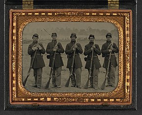 A sepia toned photograph of five soldiers standing at parade rest in a neat line