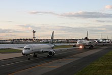 Five jumbo airplanes wait in a line on a runway next to a small body of water at John F. Kennedy Airport.