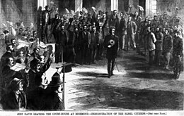 man facing forward surrounded by people on left, right and behind cheering and waving hats or handkerchiefs