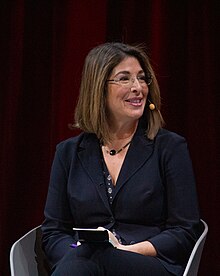 A white woman in her late 40s, dressed professionally, sits on a white chair, smiling slightly and looking to her left. She has shoulder-length dark brown hair, rimless glasses, neutral professional makeup, and a headset microphone. The background is dark red