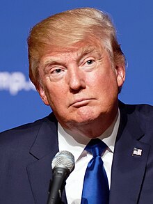 Trump at lectern before backdrop with elements of logo "TRUMP DonaldJTrump.com"