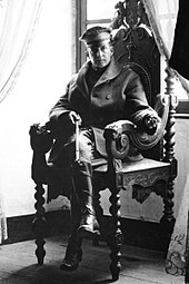 A man sits in an ornate chair. He is wearing a peaked cap, greatcoat and riding boots and holding a riding crop.