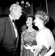 Monochrome photo of two women and a man dressed in formal attire - the two women (standing right) are smiling up at the man (facing opposite), who looks slightly amused.