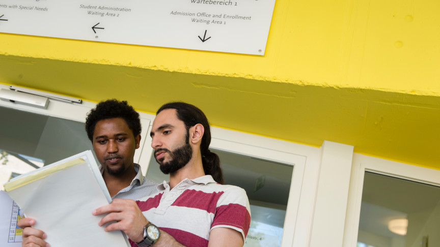 Two students stand in front of the university's enrollment office, leafing through forms together.