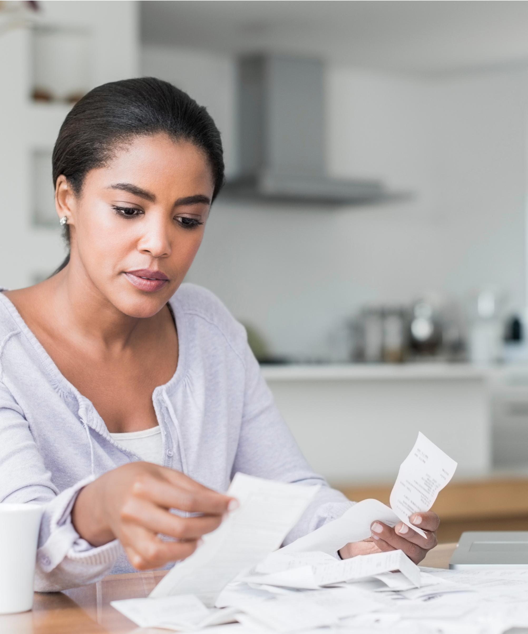 A woman sits at a table and sorts through a pile of receipts. She has a laptop to the left of the pile and a mug on the right.