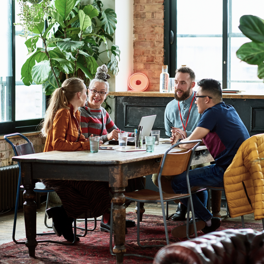 Vier Arbeitskolleg*innen sitzen an einem Besprechungstisch in einem h�bschen B�roraum mit Gr�npflanzen.