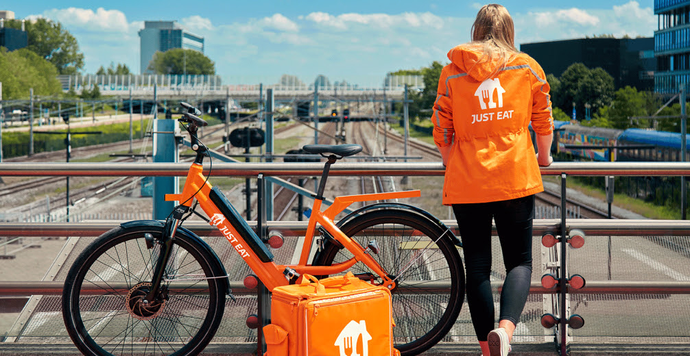 A Just Eat Takeaway.com cyclist stands next to her bike, on a bridge.